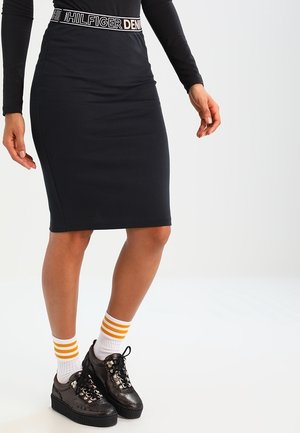 Femme portant une jupe noire cintrée avec une ceinture marquée, des chaussettes blanches à rayures orange et des chaussures à plateforme noires à lacets.