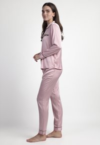 Conjunto de pijama de satén rosa que incluye una camiseta de manga larga con cuello y el logo "Barbie". Los pantalones tienen un diseño ajustado y detalles en negro en los puños.