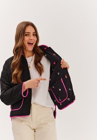 Čierna prešívaná bunda s ružovými akcentmi, ktorá obsahuje vyšívaný vnútorný podšívku s kvetinovými a postavovými dizajnmi, zipsový uzáver a vrecká.