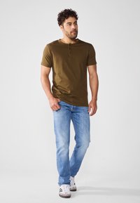 Cămașă henley cu mânecă scurtă de culoare verde măsliniu, asociată cu blugi bleu deschis și adidași gri. Cămașa are trei nasturi la guler.