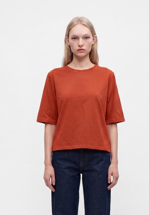 Junge Frau mit glatten blonden Haaren, die ein oranges kurzärmeliges Shirt und dunkelblaue Jeans trägt, steht vor einem einfarbigen weißen Hintergrund.