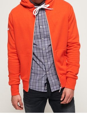 Mann trägt eine orangefarbene Kapuzenjacke mit Reißverschluss über einem schwarz-weiß karierten Hemd und dunklen Jeans, die Hände entspannt an den Seiten.