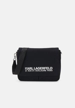 Črna torba za sporočila iz najlona, ki ima zaponko na pokrovu, nastavljiv trak in belo tiskano besedilo logotipa na sprednji strani. Minimalistična zasnova.