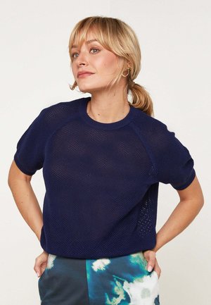 Blonde vrouw met lage paardenstaart, met gouden ronde oorbellen, een marineblauw mesh top met korte mouwen en blauw-groene broek met abstract patroon.