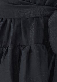 Musta mekko, jossa on teksturoitu kangas hienovaraisella kiillolla, kootut vyötärölle ja kerroksittain laskostetut hameet virtaavaa siluettia varten.
