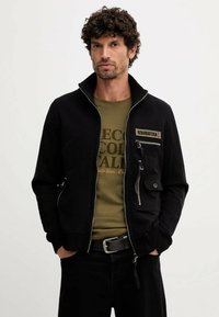 Giacca nera con zip e colletto alto, tessuto texture e molteplici tasche. Indossata sopra una maglietta grafica verde oliva.