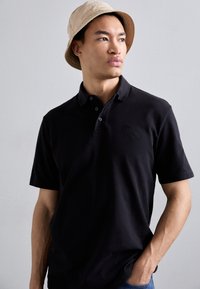 Fekete polo ing rövid ujjakkal, gallérral és két gombbal, egy világos színű kalapban lévő modellen bemutatva.