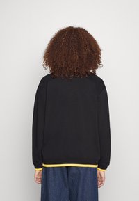 Svart sweatshirt med rund halsringning, avslappnad passform och gul trim längs nederkant och ärmar. Mjukt tyg med en texturerad yta.