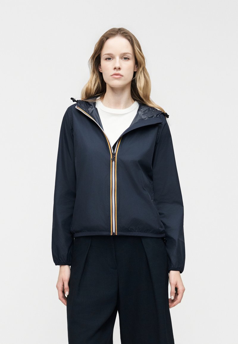 Γυναίκα με μακριά ξανθά μαλλιά φοράει μπλε ναυτικό μπουφάν με φερμουάρ και κουκούλα, λευκό πουκάμισο και σκούρο φαρδύ παντελόνι, στέκεται μπροστά σε λευκό απλό φόντο.