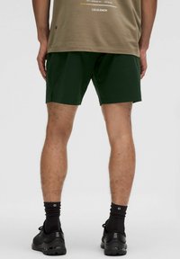 Gröna sportshorts med en slät textur, kombinerat med svarta sneakers och kontrasterande svarta ankelstrumpor. Skjortan är beige med tryckta detaljer.