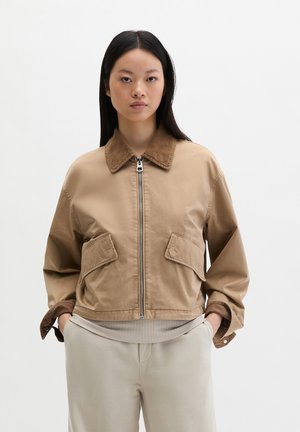 Jeune femme portant une veste beige zippée avec de grandes poches frontales et un col marron, debout avec les mains dans les poches d'un pantalon de couleur claire.