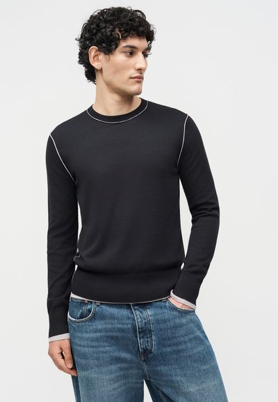 Homme aux cheveux bouclés portant un pull noir à manches longues avec des coutures blanches et un jean bleu, regardant sur le côté avec une main dans la poche.