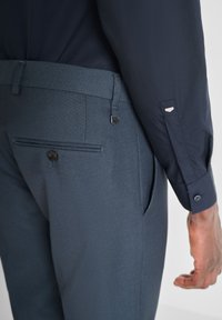 Camicia blu navy a bottoni con un pattern testurizzato, abbinata a pantaloni grigi scuri su misura con una tasca posteriore e dettaglio a pressione.