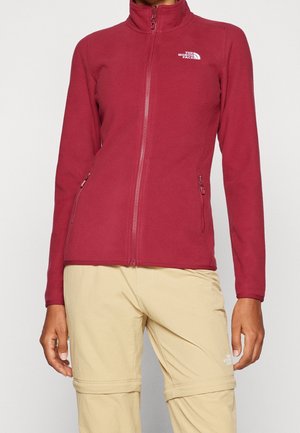 Femme portant une veste polaire rouge zippée avec des poches latérales zippées et un pantalon convertible beige. Les deux articles arborent le logo The North Face.