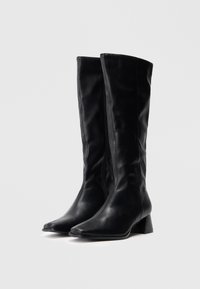 Une paire de bottes en cuir noires mi-mollet avec de petits talons carrés et des zips sur les côtés, sur un fond blanc uni.