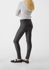 Pantalons noirs ajustés avec une texture lisse, dotés de poches arrière et d'une taille haute. Portés avec des ballerines noires et un pull gris.