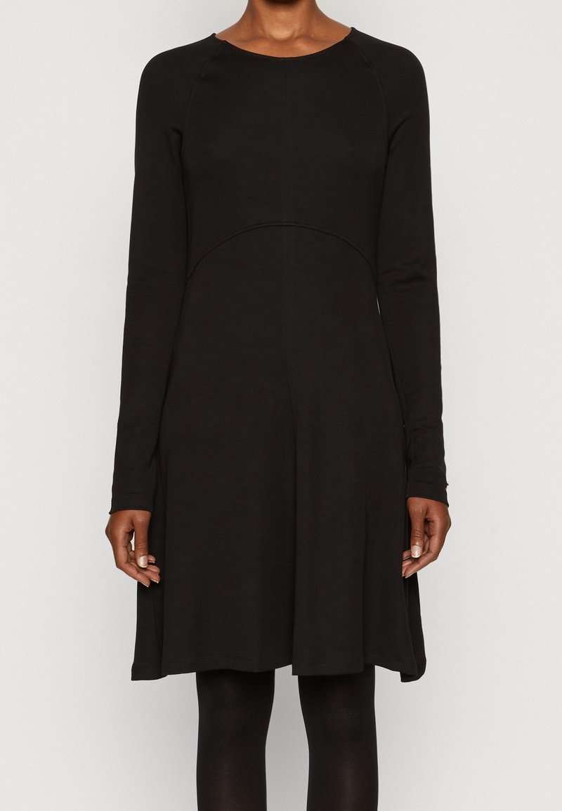 Femme portant une robe noire à manches longues avec une jupe trapèze, debout devant un fond clair uni.