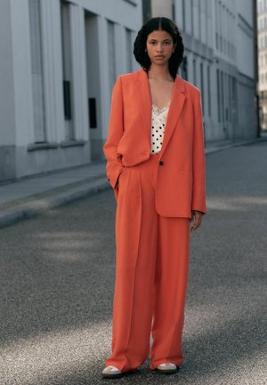 Woman standing on street wearing bright orange oversized suit with white polka-dot camisole and gray sneakers, hands in pockets.
