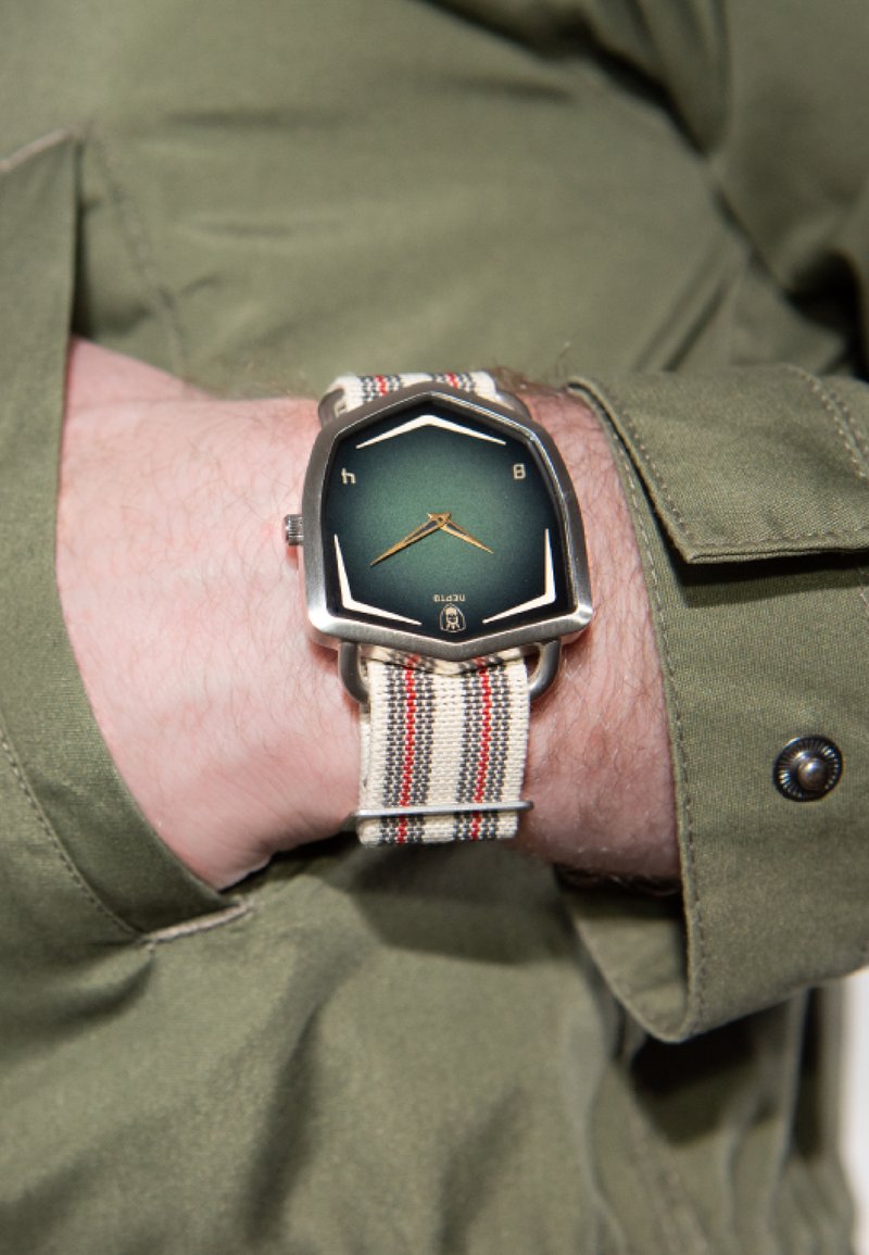 Armbåndsur med en hexagonal grøn urskive, guld hænder og et vævet beige rem med røde og sorte striber, båret over en olivengrøn skjorte.