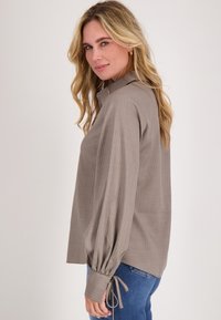 Lichtbruine gestreepte blouse met een kraag en wijdvallende mouwen met gerimpelde manchetten. Gecombineerd met blauwe jeans, zijaanzicht.