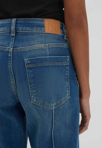 Jeans in denim blu con taglio dritto, caratterizzati da una toppa in pelle sulla vita e due tasche posteriori con dettagli di cucitura visibili.