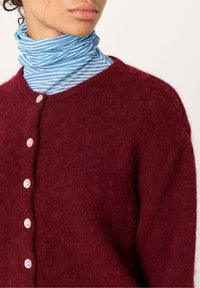 Burgundski pleteni kardigan z dlačastim teksturama, s petimi roza gumbi na sprednji strani, nosi se čez modro-beli �črtasti pulover s turtleneck ovratnikom.