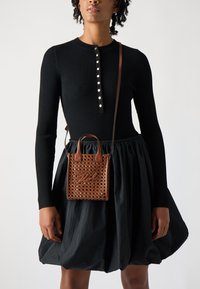 Rjava tkana torba z dvema kratkimi ročaji, z značilno teksturo in embosiranim napisom, nošena čez ramo preko črne rebraste obleke.