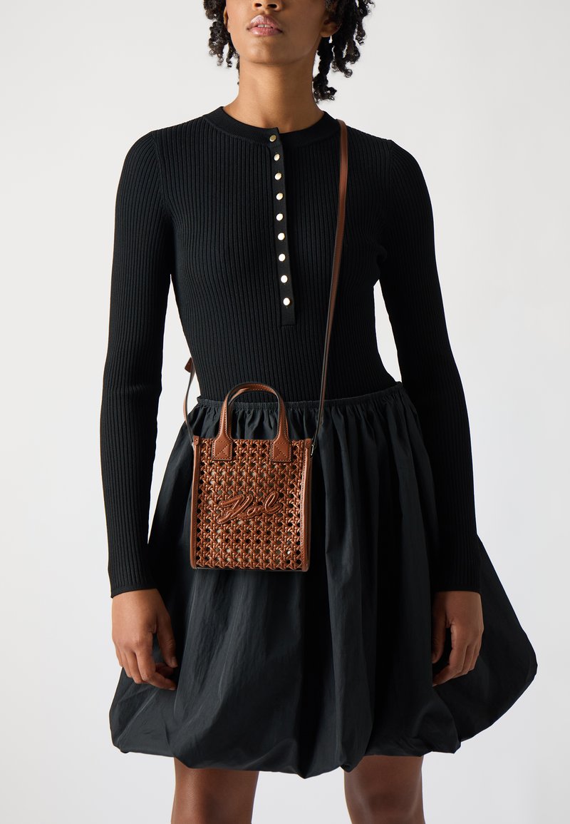 Rjava tkana torba z dvema kratkimi ročaji, z značilno teksturo in embosiranim napisom, nošena čez ramo preko črne rebraste obleke.