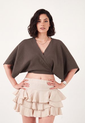 Donna con capelli corti e ricci che indossa una maglietta corta marrone e ampia e una minigonna beige a balze sovrapposte davanti a uno sfondo bianco semplice.