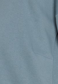 Světle modrá mikina s jemnou texturou, kulatým výstřihem a přední kapsou. Tkanina vypadá hladce s jemnými detaily šití.