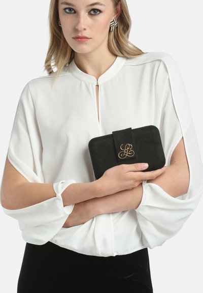Fekete velúr clutch arany díszcsattal, egy fehér blúz mellett, amelynek lágy, folyamatos ujjai vannak. Simaság és elegáns dizájn jellemzi.