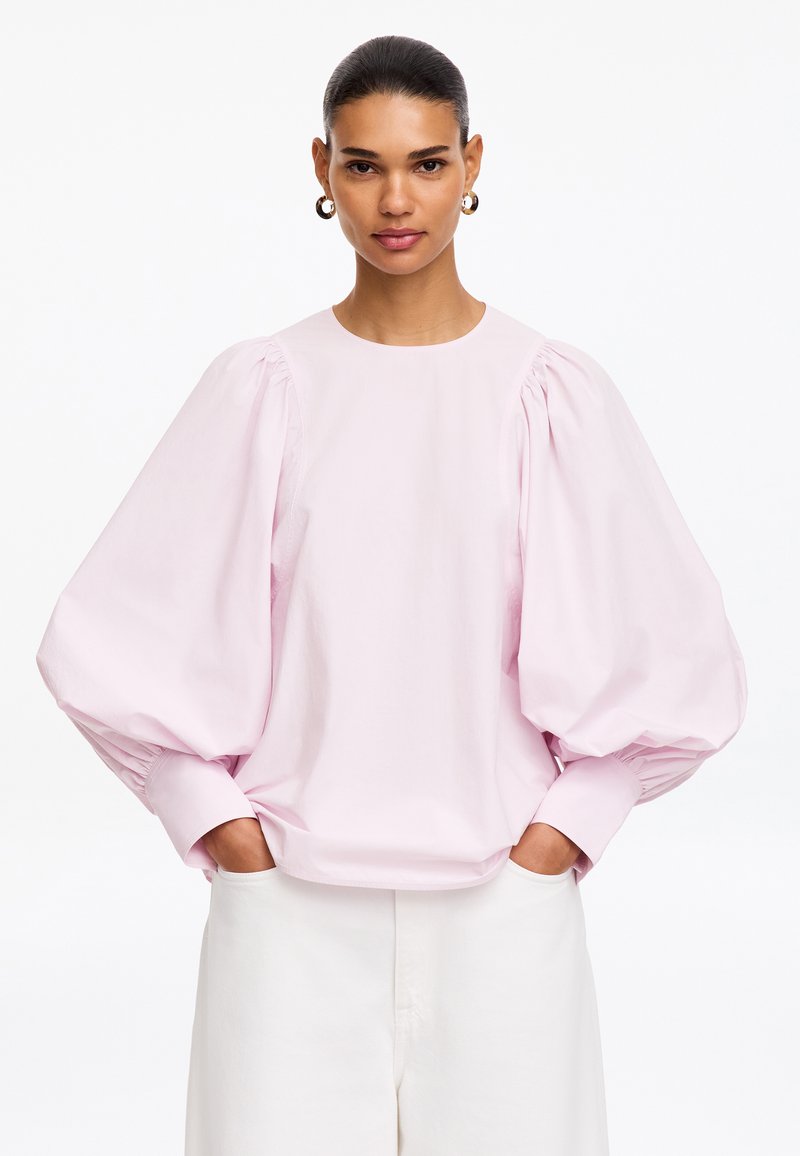 Femme portant un chemisier rose pâle à grandes manches bouffantes et un pantalon blanc, debout les mains dans les poches sur fond blanc.