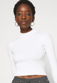 Top court à manches longues blanc côtelé avec un col montant. Présente un tissu texturé et une coupe ajustée. Porté avec un pantalon taille haute à motif.