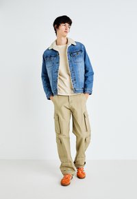 Jeansjacke mit einem Schafsfellkragen, beigem Langarmhemd, Cargo-Hosen und orangenen Turnschuhen. Das Outfit bietet ein lässiges, geschichtetes Erscheinungsbild.