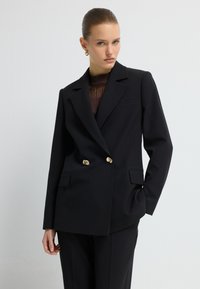 Czarna dwurzędowa marynarka z złotymi guzikami, o dopasowanym kroju, z wyraźnie zarysowanymi ramionami i dwoma przednimi kieszeniami. Gładka tekstura materiału.