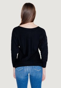 Maglione nero a maniche lunghe con scollatura ampia, texture liscia e scritte ricamate sulla manica, abbinato a jeans in denim blu.