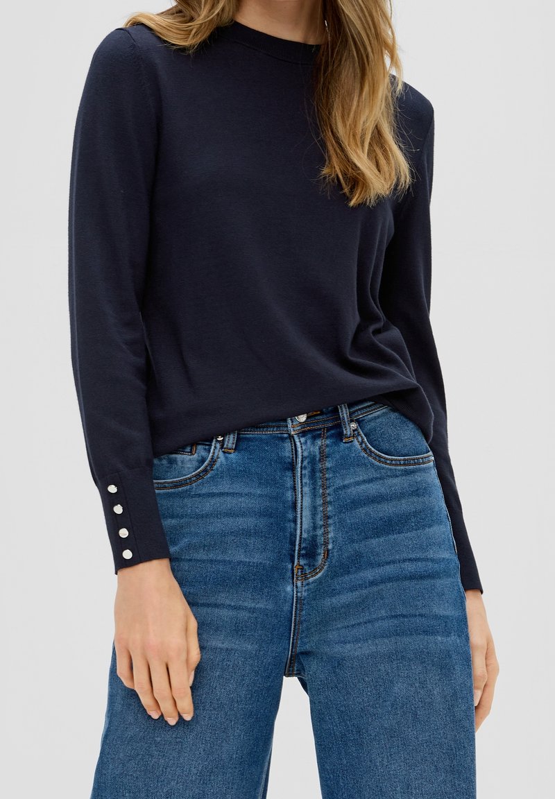 Pull en maille bleu marine à manches longues, avec des boutons décoratifs aux poignets. Porté avec un jean taille haute bleu clair.