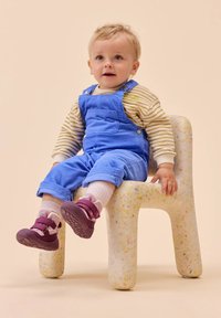 Salopette blu in corduroy, camicia a maniche lunghe a righe, calzini rosa e scarpe viola su un bambino seduto su una sedia a trama punteggiata.