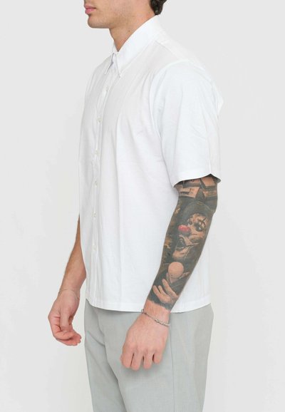 Camicia button-up a maniche corte in tessuto di cotone bianco, caratterizzata da un colletto classico e un orlo dritto. Il modello indossa pantaloni grigi chiari.