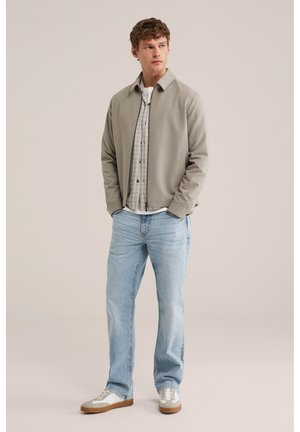 Jauns vīrietis stāv, valkājot gaiši pelēku rāvējslēdzēja jaku pār kreklu ar rūtiņu rakstu, gaiši zili džinsi un baltus sporta apavus ar gumijas zolēm.