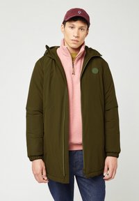Olijfgroene pufferjas met een ritssluiting aan de voorkant en een capuchon, gedragen over een lichtroze fleece vest met ritssluiting. Model draagt ook een bordeauxrode pet.