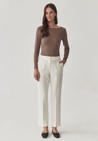 Braunes langärmeliges Oberteil und cremefarbene, taillierte Hose mit Falten. Schwarze Ballerinas runden das Outfit ab. Schlichter Hintergrund, der sich auf die Details der Kleidung konzentriert.