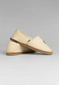 Espadrilles de color beige claro con puntera redondeada, suela de yute trenzado y detalle del logo en negro. Parte superior de tela suave, diseño plano para uso casual.