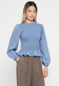 Lys blå smockbluse med lange puffærmer og en bølget kant, lavet af tekstureret stof. Sammen med højtaljede brune bukser.