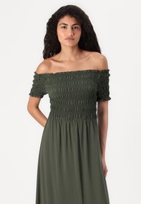 Kvinna med långt lockigt hår klädd i en olivgrön off-shoulder-klänning med rynkad kropp, stående mot en enkel vit bakgrund.