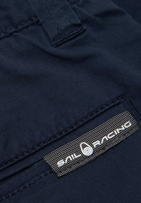 Navy sininen kangas, jossa on sileä pinta. Varustettu ommellulla tarralla, jossa lukee "SAIL RACING" valkoisella tekstillä sekä hieman kohotettu pyöreä logo.