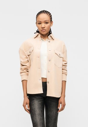 Jeune femme aux cheveux tressés portant une surchemise en velours côtelé beige, un haut blanc raccourci boutonné et un pantalon noir texturé, se tenant devant un fond blanc.