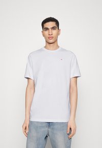 T-shirt bianca in cotone con scollatura rotonda, maniche corte e un piccolo logo rosso sul petto. Indossata con jeans denim blu chiaro.