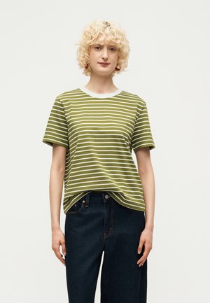 Άτομο με κοντά σγουρά ξανθά μαλλιά που φοράει πράσινο και λευκό ριγέ μπλουζάκι, μπαλωμένο μέσα σε σκούρο μπλε τζιν, στέκεται μπροστά σε ένα απλό φωτεινό φόντο.