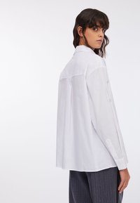 Chemise blanche à manches longues avec une coupe droite, des poignets boutonnés et des coutures décoratives le long des manches et du dos. Tissu doux au toucher.